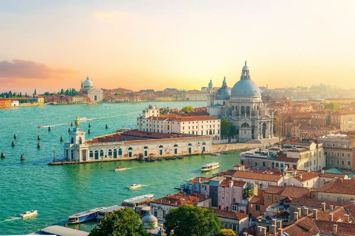 fine-vintage-ltd-veneto-tour-venice-1200x800 An expansive view shows the city of Venice, Italy from above. Domes and buildings with red tile roofs stretch across the frame, with bodies of water on either side.