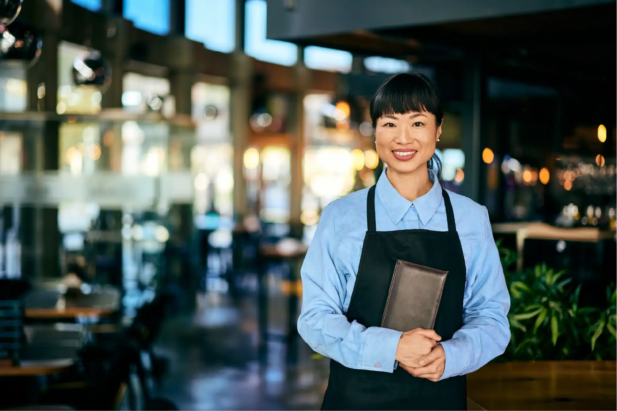 A smiling restaurant server in an apron holding a menu in a warmly lit bar.